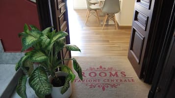 En hotellentrén med en krukväxt, en dörrmatta med texten ”ROOMS” och ”TAZIONE CENTRALE”, och en matplats i bakgrunden.