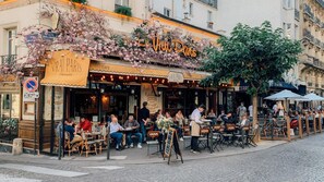 An outdoor café with tables and chairs, a sign reading 'Le Vie Dan's,' and a street view with buildings in the background. - The Montmartre 1881 (Paris)
