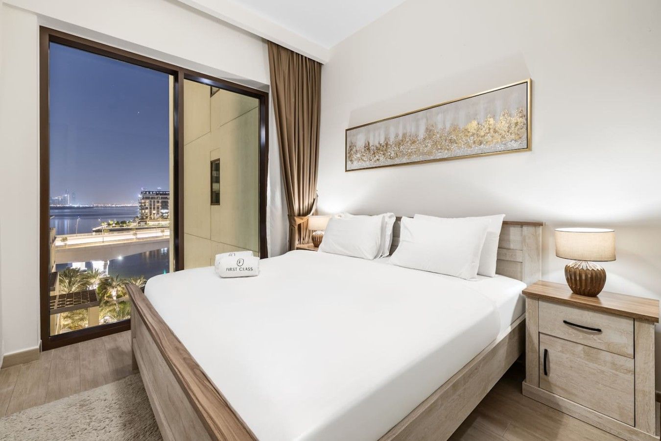 A hotel room with a large bed, a nightstand, a lamp, a painting, and a view of a cityscape through a sliding door.