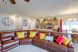 A modern living room with a brown leather sectional sofa, a large wall clock, and a kitchen area with wooden cabinets and stainless steel appliances.