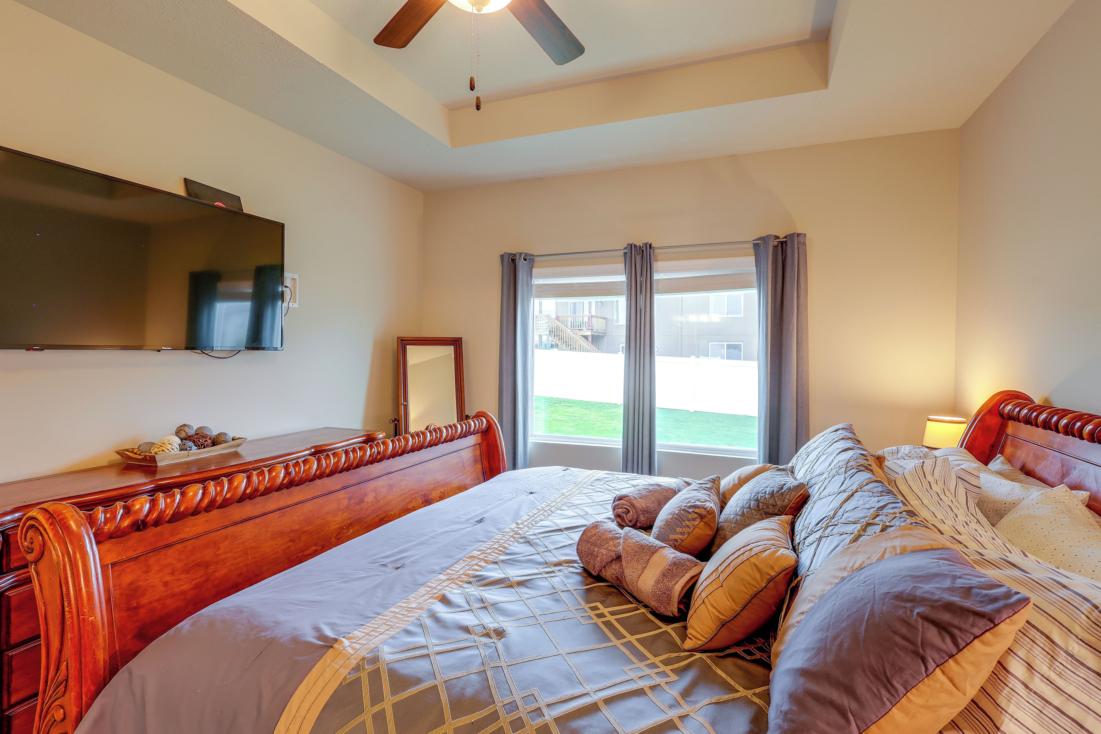 A bedroom with a large bed, a flat-screen TV, a ceiling fan, and a window with curtains.