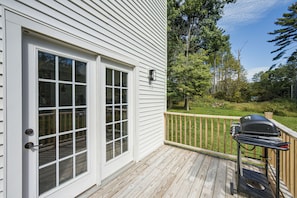 A wooden deck with a barbecue grill, a white door with glass panels, and a view of a green landscape with trees.