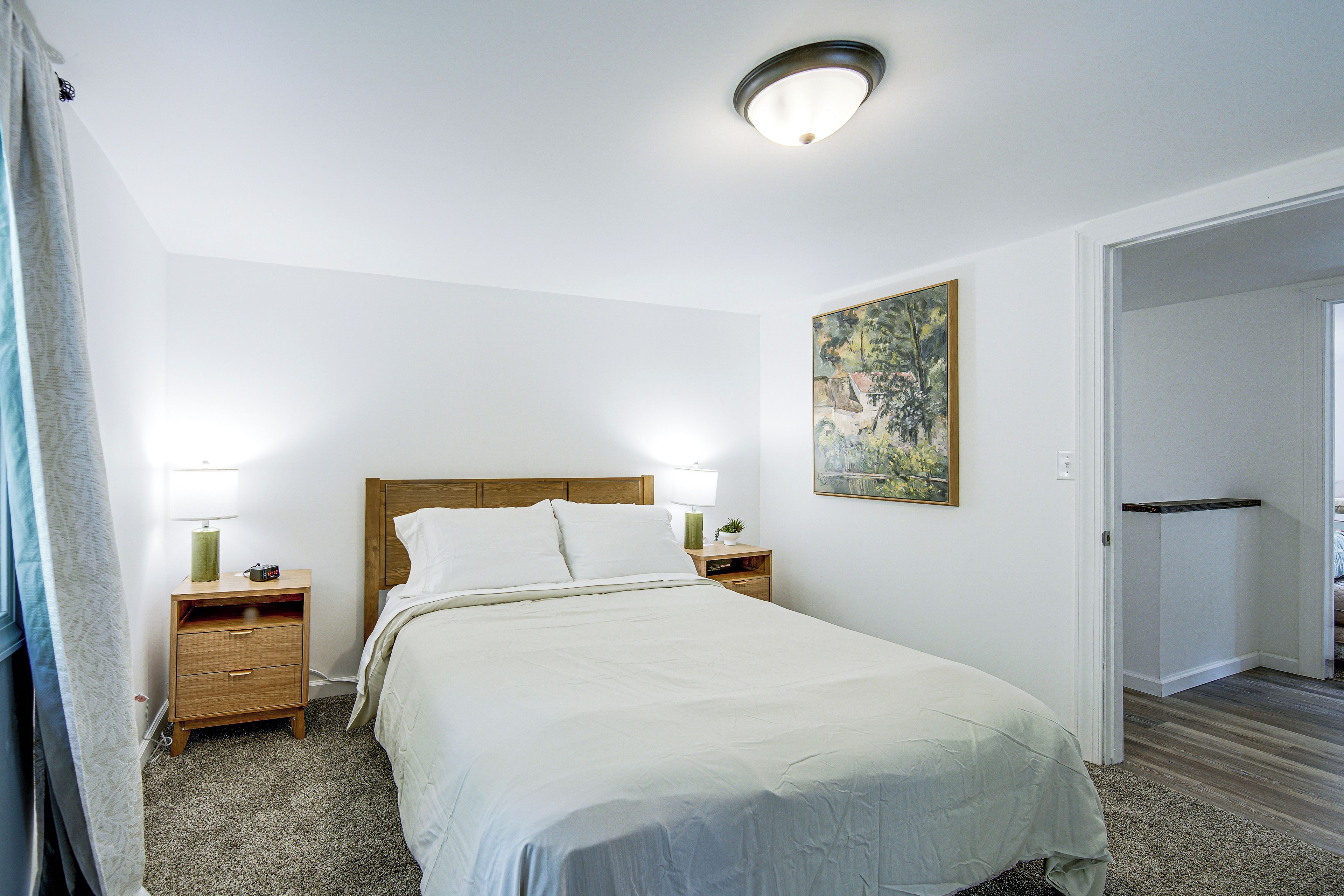 Un dormitorio con cama, dos mesitas de noche, una mesita auxiliar, un cuadro en la pared y una puerta que da a otra habitación.