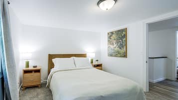 Un dormitorio con cama, dos mesitas de noche, una mesita auxiliar, un cuadro en la pared y una puerta que da a otra habitación.