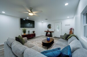 A living room with a large sectional sofa, a coffee table, a flat-screen TV mounted on the wall, a ceiling fan, and a decorative mirror.