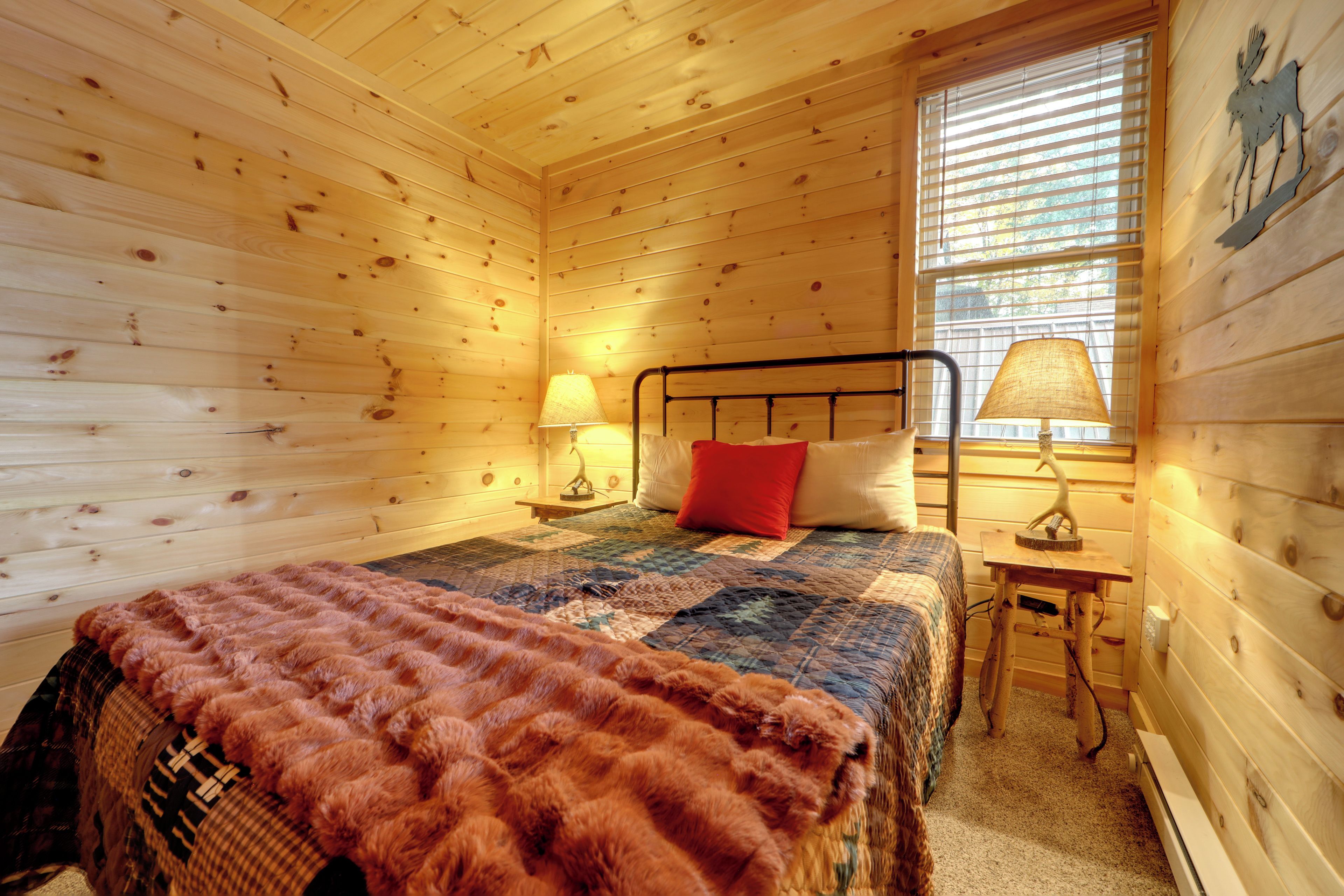 A cozy wooden cabin bedroom with a bed, two bedside lamps, a window with blinds, and a wooden headboard.