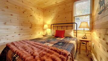 A cozy wooden cabin bedroom with a bed, two bedside lamps, a window with blinds, and a wooden headboard.