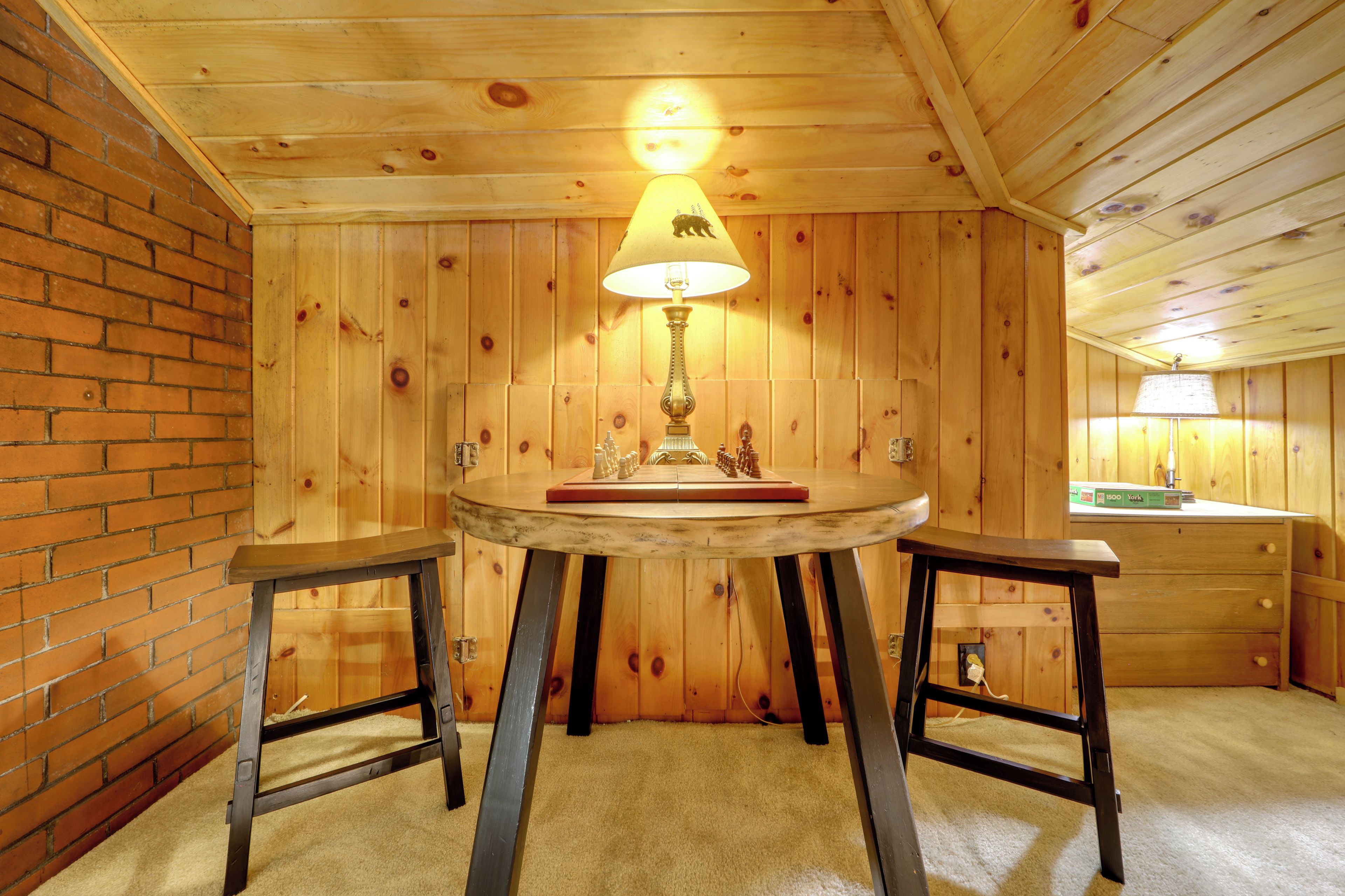 A wooden room with a table and two stools, a lamp, and a brick wall.