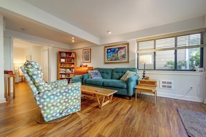 A living room with a sofa, a chair, a coffee table, a bookshelf, and a painting on the wall. - Walk to Downtown Edmonds! Condo w/ Patio (Edmonds)