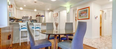 A modern kitchen with white cabinets, a stainless steel microwave, and a refrigerator. There is a dining area with a glass table and blue upholstered chairs.