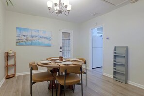 Un coin repas avec une table ronde, des chaises et un lustre. Un tableau orne le mur, et une porte donne accès à une autre pièce.