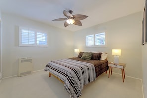 Une chambre à coucher comprenant un lit, deux lampes de chevet, un ventilateur de plafond et deux fenêtres équipées de stores.