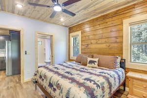 Un dormitorio con techo de madera, una cama con colcha floral, una mesita de noche y un ventanal con vista a árboles.