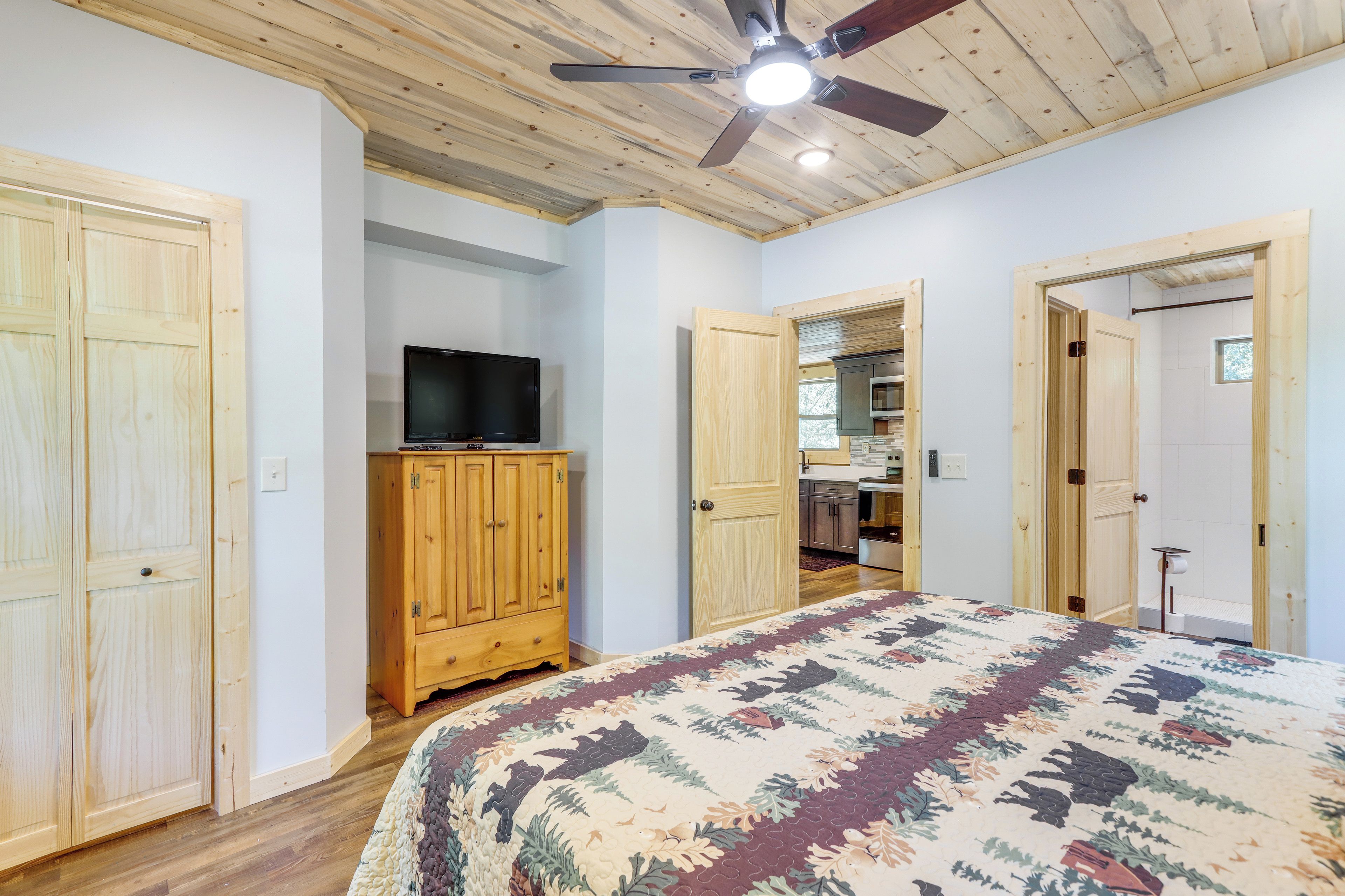 Un dormitorio con cama, armario de madera, televisión y ventilador de techo, con vistas a una zona de cocina.