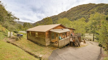 A wooden cabin with a deck, surrounded by a forested hillside.