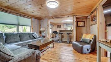 A cozy cabin interior with a wooden ceiling, a sofa, a dining table, a leather chair, and a kitchen area.