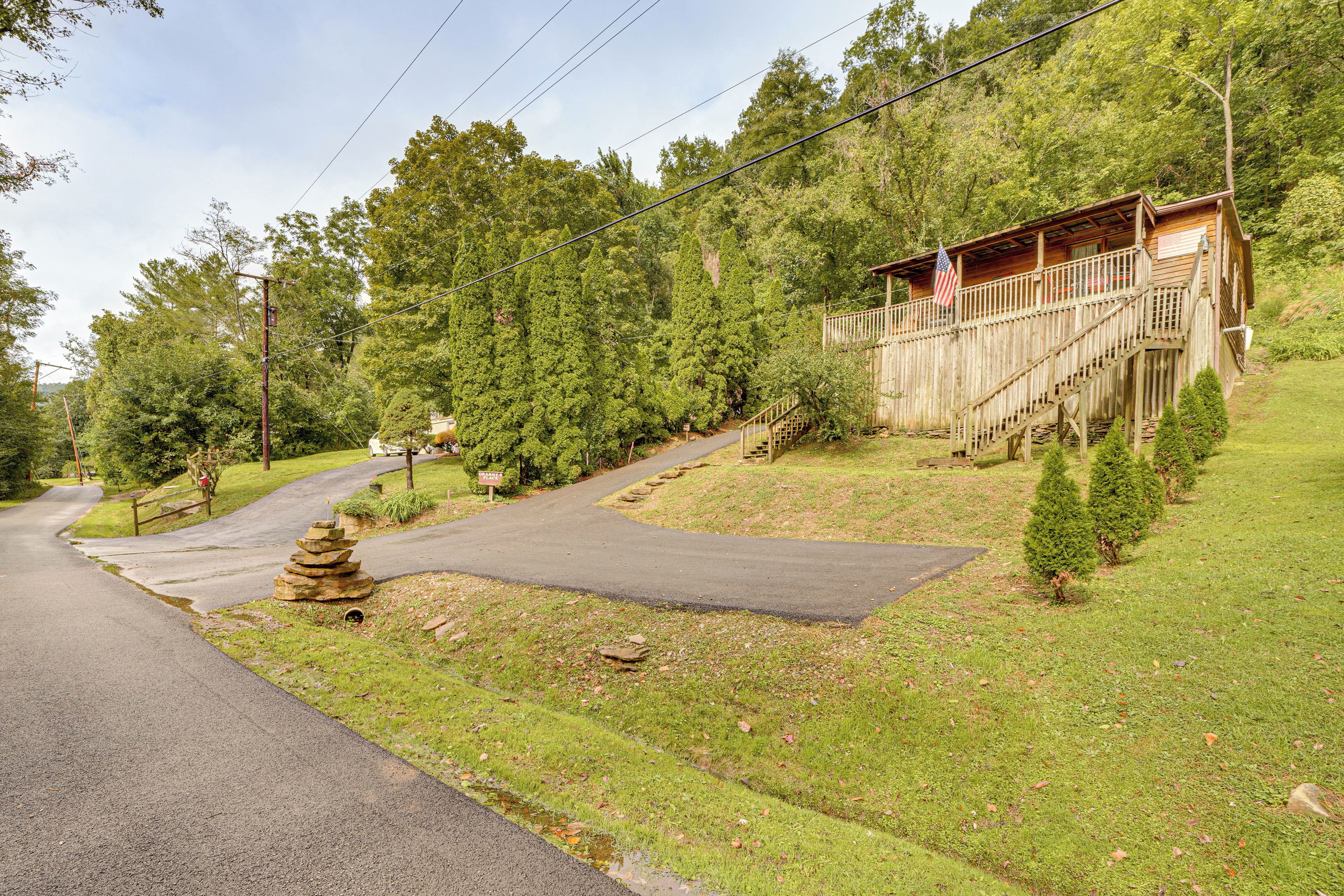 A wooden cabin with a porch and a flag, surrounded by trees and a paved driveway.