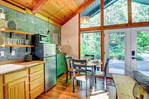 Cocina compacta con refrigerador, microondas y gabinetes de madera. Cuenta con una zona de comedor con mesa y sillas, y un amplio ventanal con vistas al exterior.