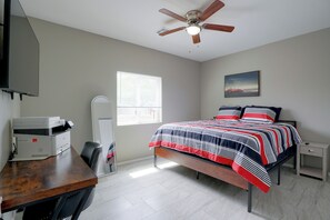Ein Schlafzimmer mit einem Bett, einem Schreibtisch mit Drucker, einem Deckenventilator, einem Fenster und einem Bild an der Wand.