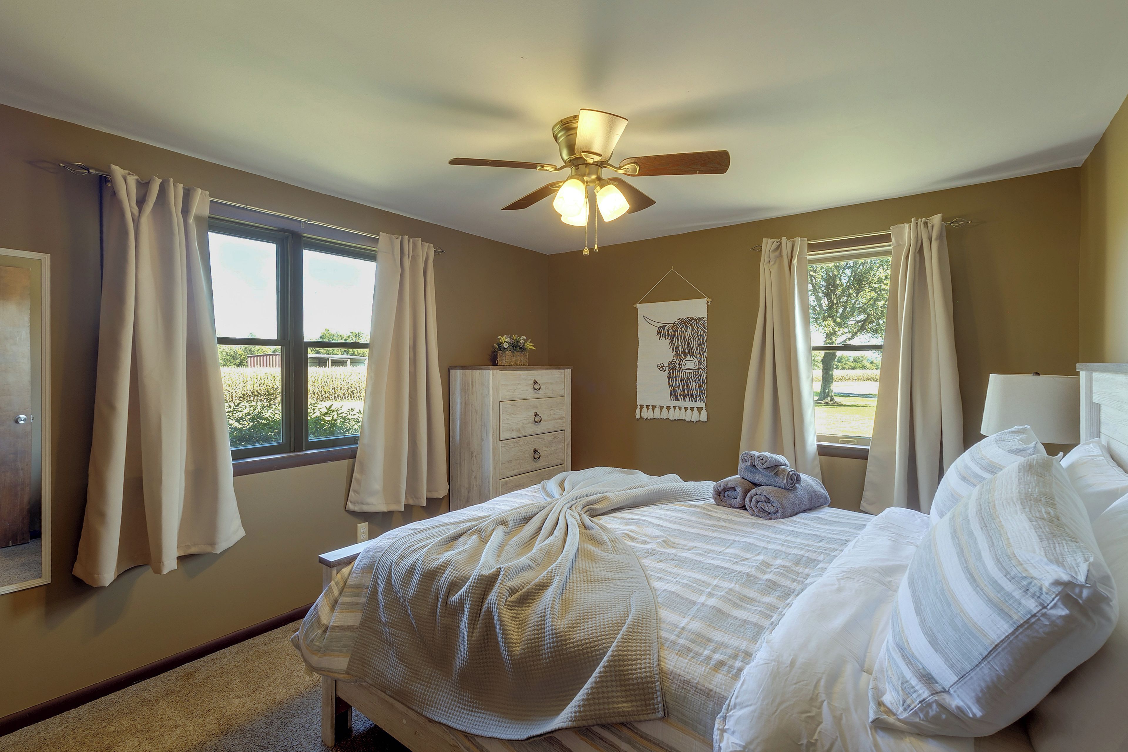 Un dormitorio con cama, cómoda, ventilador de techo y ventanas con cortinas.