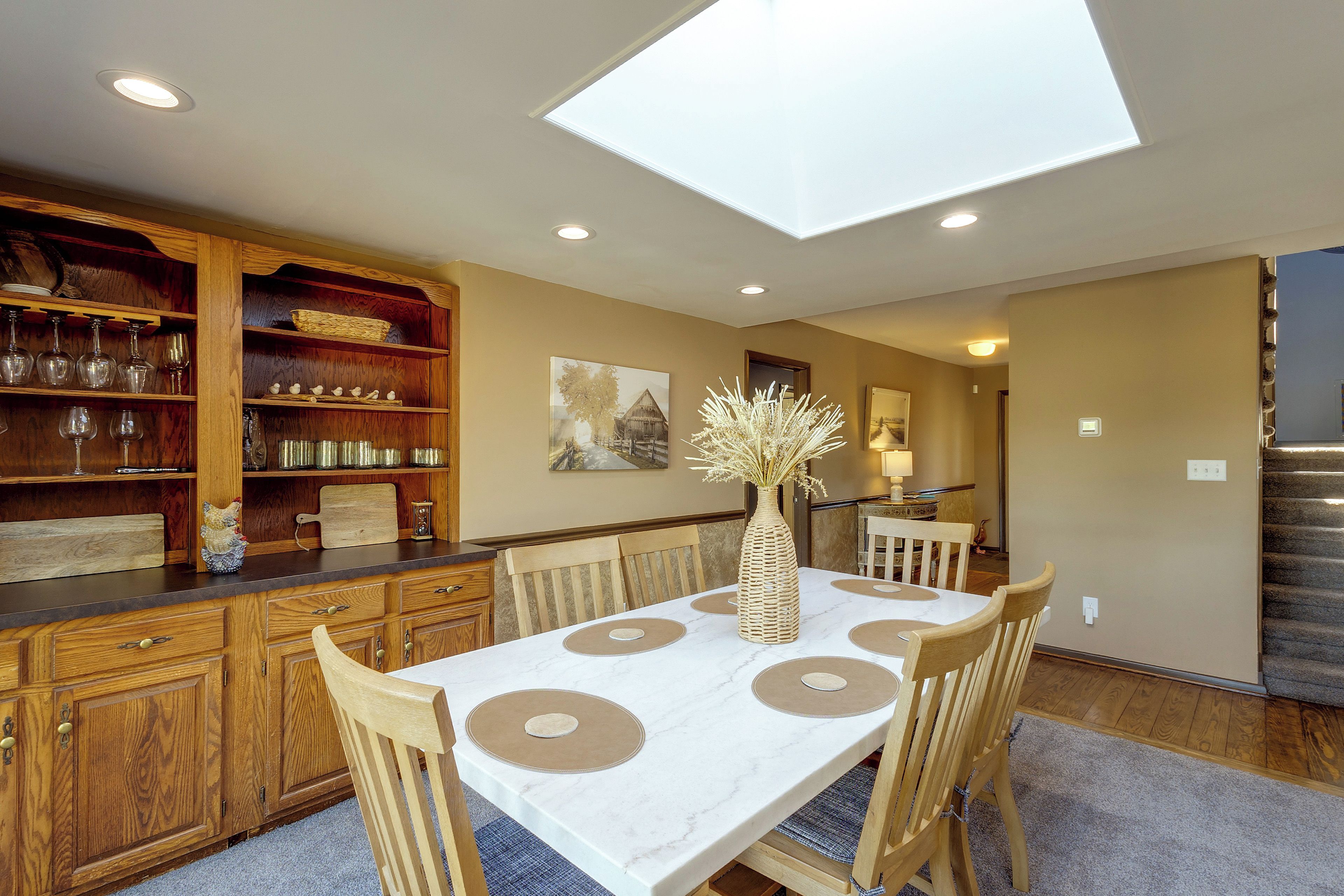 A dining area with a table set for four, surrounded by wooden chairs, a wooden buffet with glassware, and a skylight above.