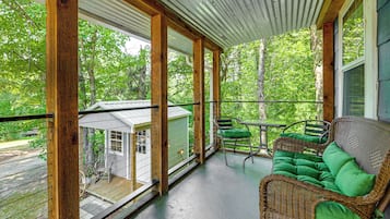 Une véranda couverte avec un canapé et des fauteuils en osier, une petite table et une vue sur une maison et des arbres.