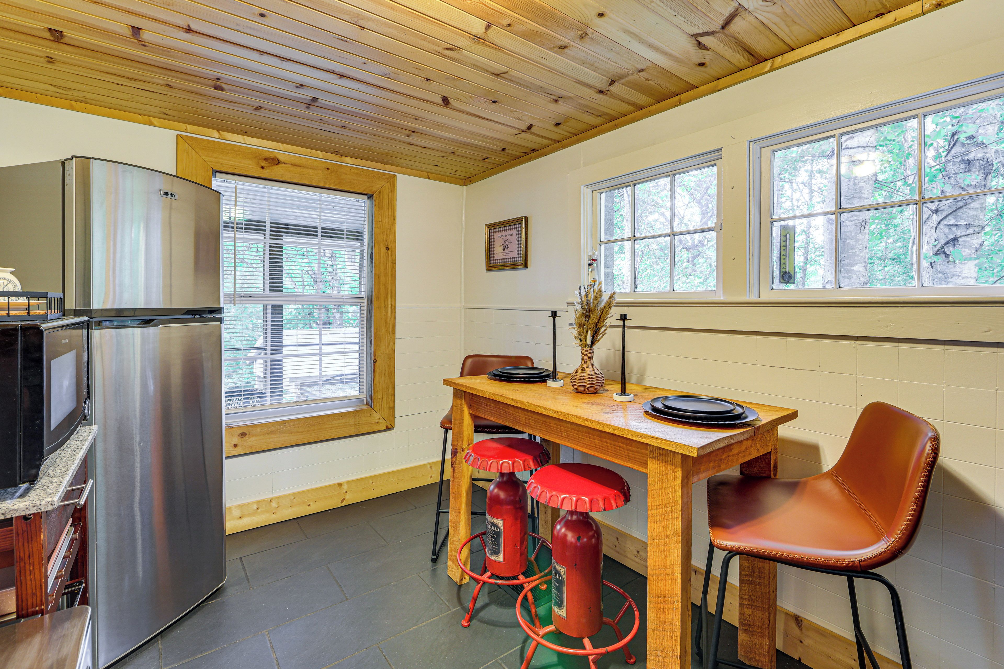 A compact kitchen with a stainless steel refrigerator, a wooden dining table with two red stools, and a window with a view of trees.