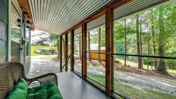 Une véranda couverte, avec un fauteuil en osier et un coussin vert, donnant sur une zone boisée avec un chemin de gravier et des véhicules garés.