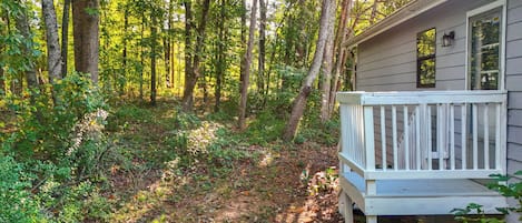 A cabin with a deck, surrounded by a forest with tall trees and a ground covered in leaves.