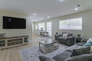 A modern living room with a flat-screen TV, a grey sofa, a coffee table, and a dining area with a table and chairs.