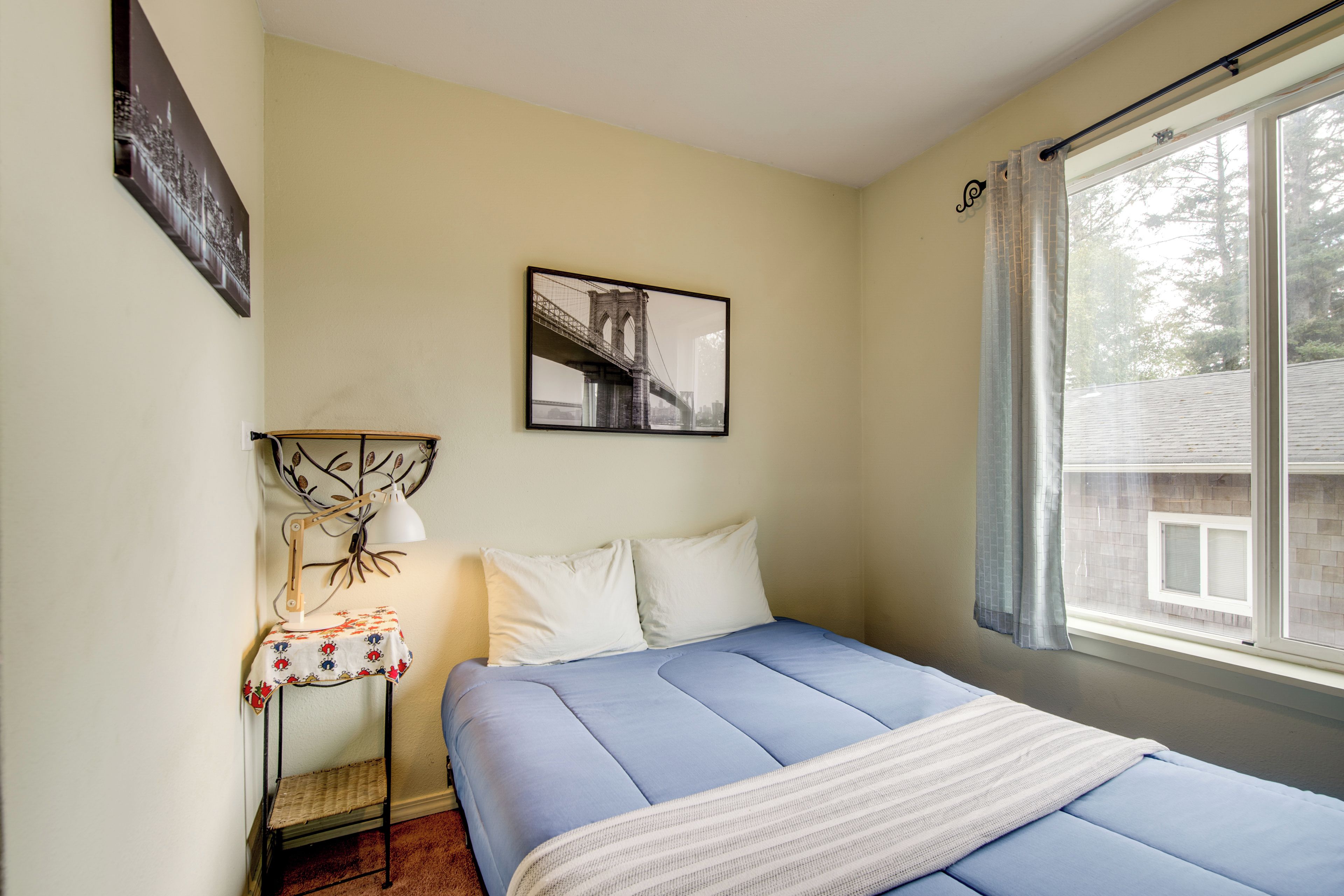 A bedroom with a bed, a bedside table with a lamp, a framed picture on the wall, and a window with curtains.