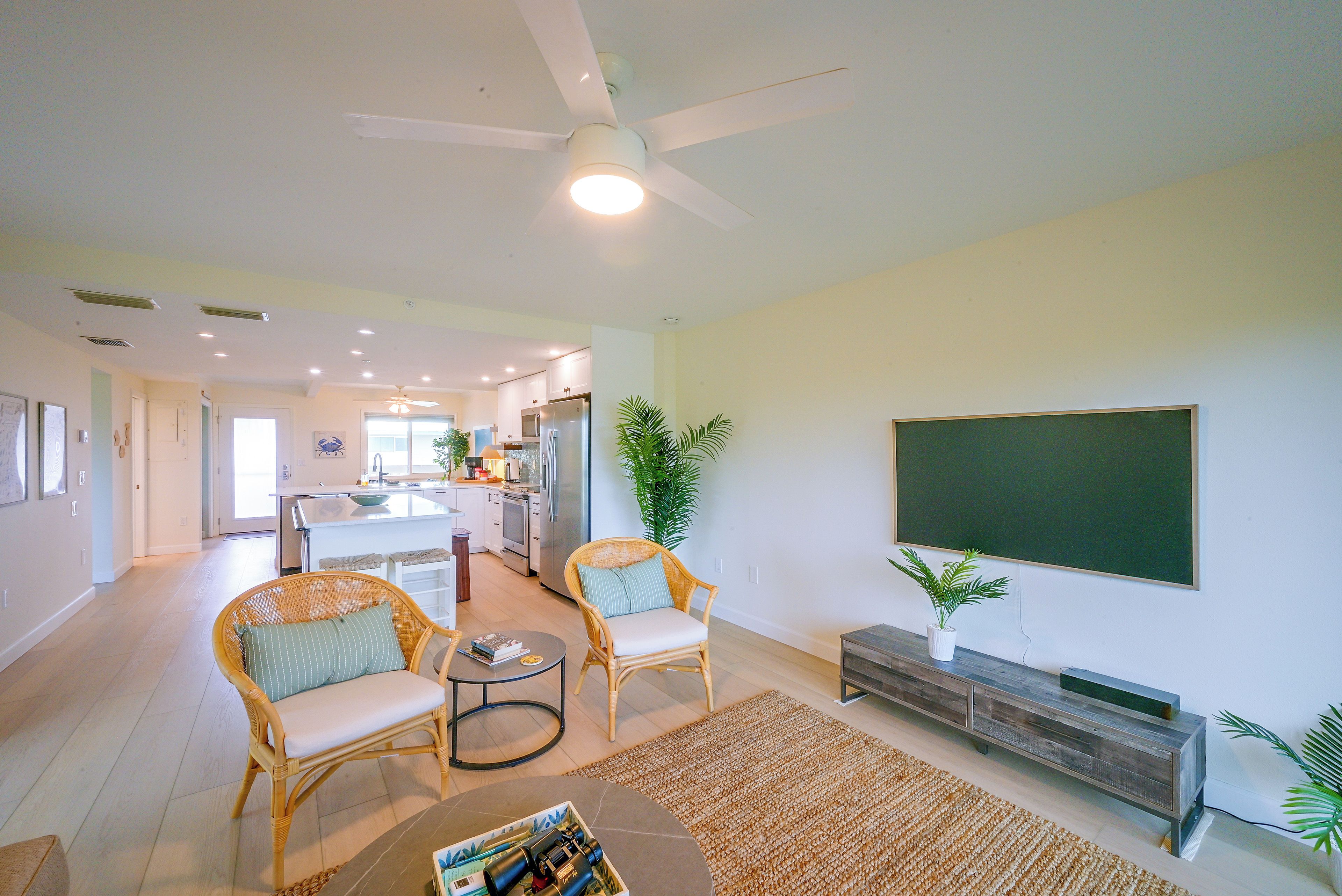 A spacious living room with a large flat-screen TV, comfortable seating, a coffee table, and a kitchen area in the background.