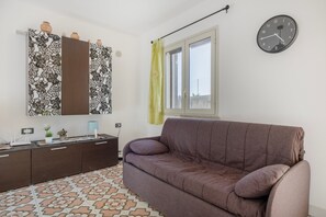 A living room with a brown sofa, a patterned cabinet, a wall clock, and a window with a curtain. - Apartment 'Granchio Blu Porto Palo di Menfi' with sea view and Wi-Fi (Porto Palo Est)