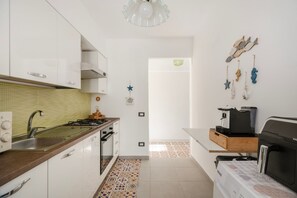 A kitchen with white cabinets, a gas stove, a coffee machine, and a wall-mounted rack with hanging items. - Apartment 'Granchio Blu Porto Palo di Menfi' with sea view and Wi-Fi (Porto Palo Est)