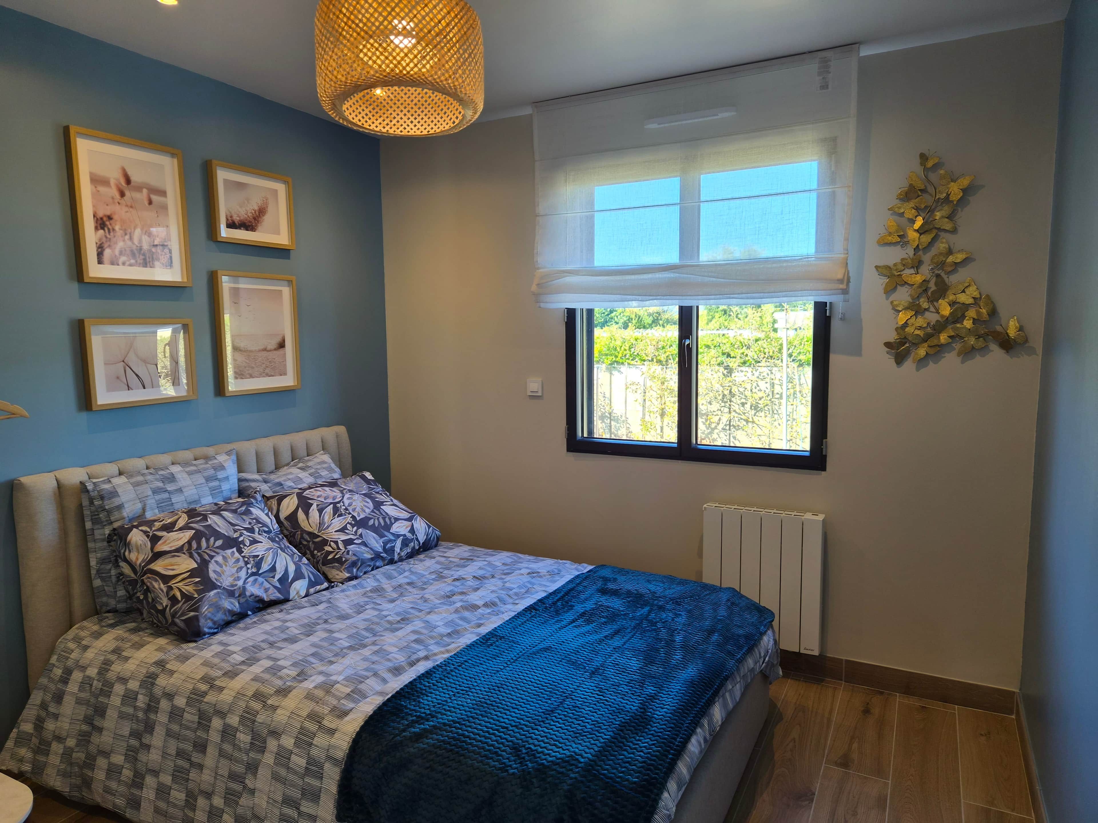 Une chambre à coucher avec un lit, des tableaux encadrés accrochés au mur, une fenêtre donnant sur un paysage et un radiateur.