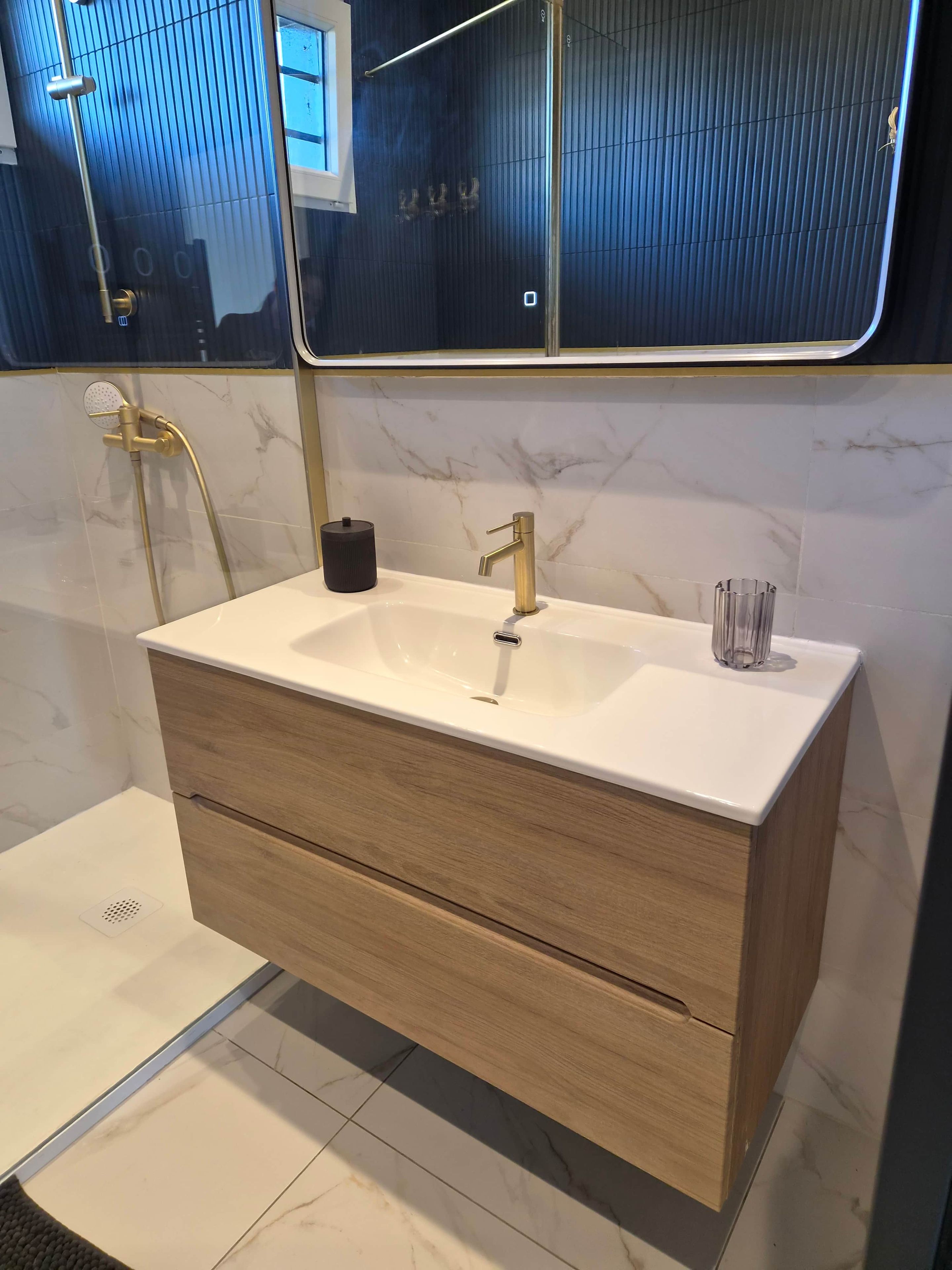 Une salle de bain avec un lavabo blanc, des robinetteries dorées, un meuble-lavabo en bois et un miroir qui reflète la pièce.