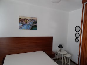 Et hotelværelse med en seng, et lille bord med en lampe, et billede af en bro på væggen og en dør med gardin.