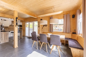 Ein modernes Interieur mit Holzbalkendecke, einem Essbereich mit grau gepolsterten Stühlen und einer Küche mit Edelstahlgeräten.