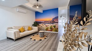 Un salon moderne avec un canapé, une table basse ronde et une fresque murale représentant un coucher de soleil sur une ville côtière.