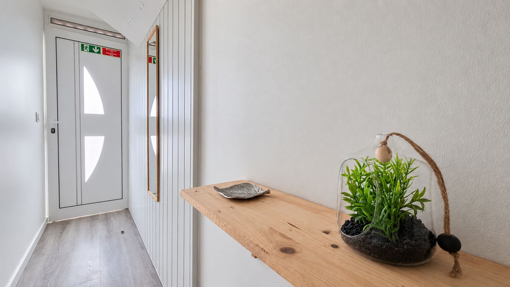 Un couloir avec une porte blanche, une étagère en bois ornée d’une plante et une petite assiette.