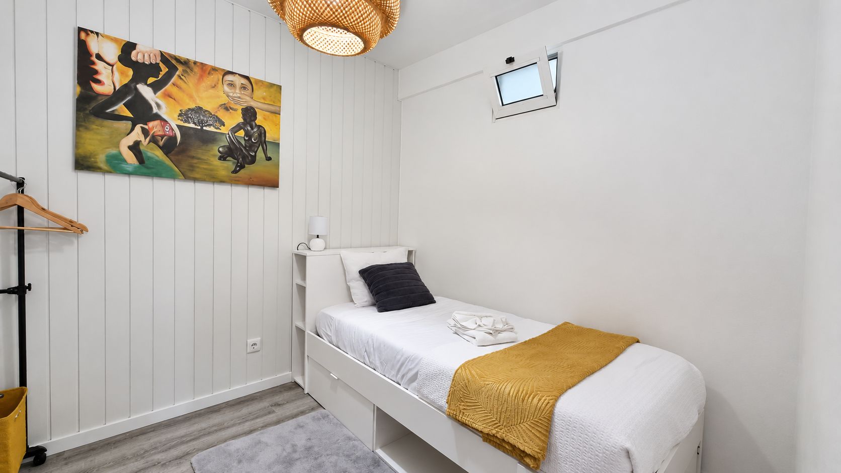 Een enkel bed met een wit hoofdeinde, een zwart kussen en een mosterdkleurige deken. Een klein wit nachtkastje met een lamp staat links van het bed. Een schilderij met abstracte figuren hangt aan de muur boven het bed.