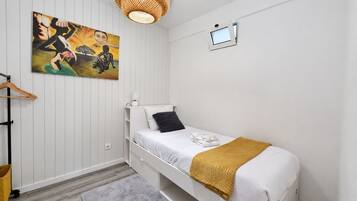 A single bed with a white headboard, a black pillow, and a mustard-colored throw blanket. A small white nightstand with a lamp is to the left of the bed. A painting with abstract figures is on the wall above the bed.