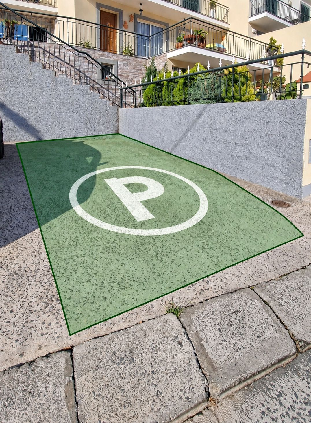 Un estacionamiento con superficie verde y un símbolo de 'P' blanco, rodeado de escalones de piedra y un edificio con balcones.