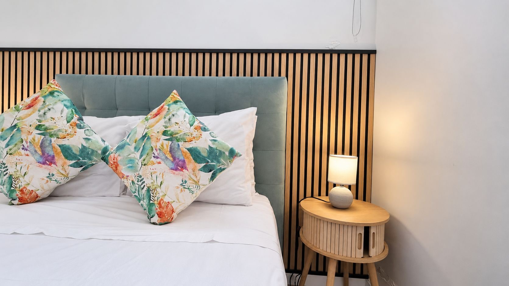 Ein Bett mit weißer Bettwäsche, zwei blumige Kissen, einem grauen Kopfteil und einem Nachttisch mit Lampe.