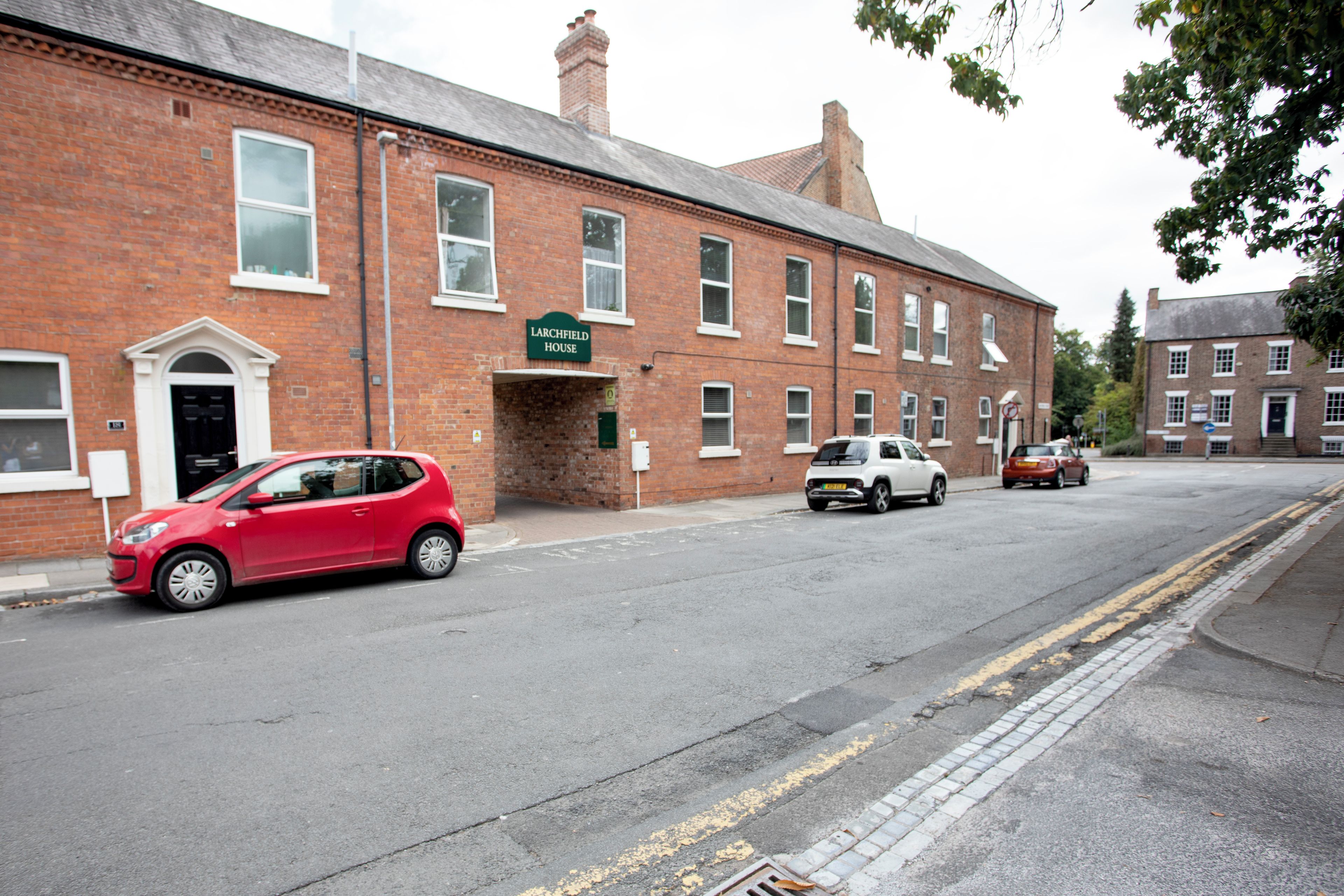 A red car parked in front of a brick building with a sign reading 'Larchfield House.'