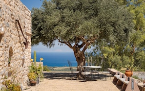 Edificio de piedra con un gran árbol en primer plano, mesas al aire libre y vista al mar.