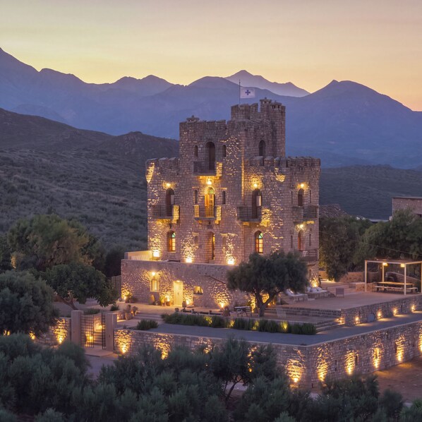 A historic stone building with multiple towers and arched windows, surrounded by a landscaped garden and a mountainous backdrop.