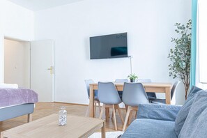 Ein modernes Wohnzimmer mit Sofa, Couchtisch, Esstisch mit Stühlen, einem Wand-Fernseher und einem Blumentopf.