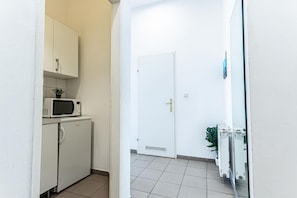 Ein schmaler Flur mit einer Mikrowelle und einem kleinen Kühlschrank links, einer weißen Tür rechts und einer Blumentopfanlage in der Nähe der Tür.