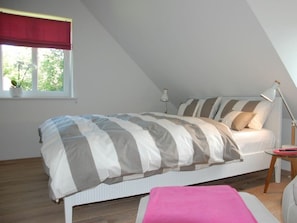 Ein Schlafzimmer mit einem Bett, einem Nachttisch, einer Lampe, einem Fenster mit Blick auf Bäume und einem roten Vorhang.